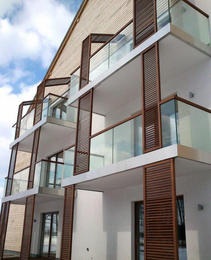 Fasada nowoczesnego budynku z balkonami z drewnianymi żaluzjami i szklanymi balustradami, widok z dołu na błękitne niebo.