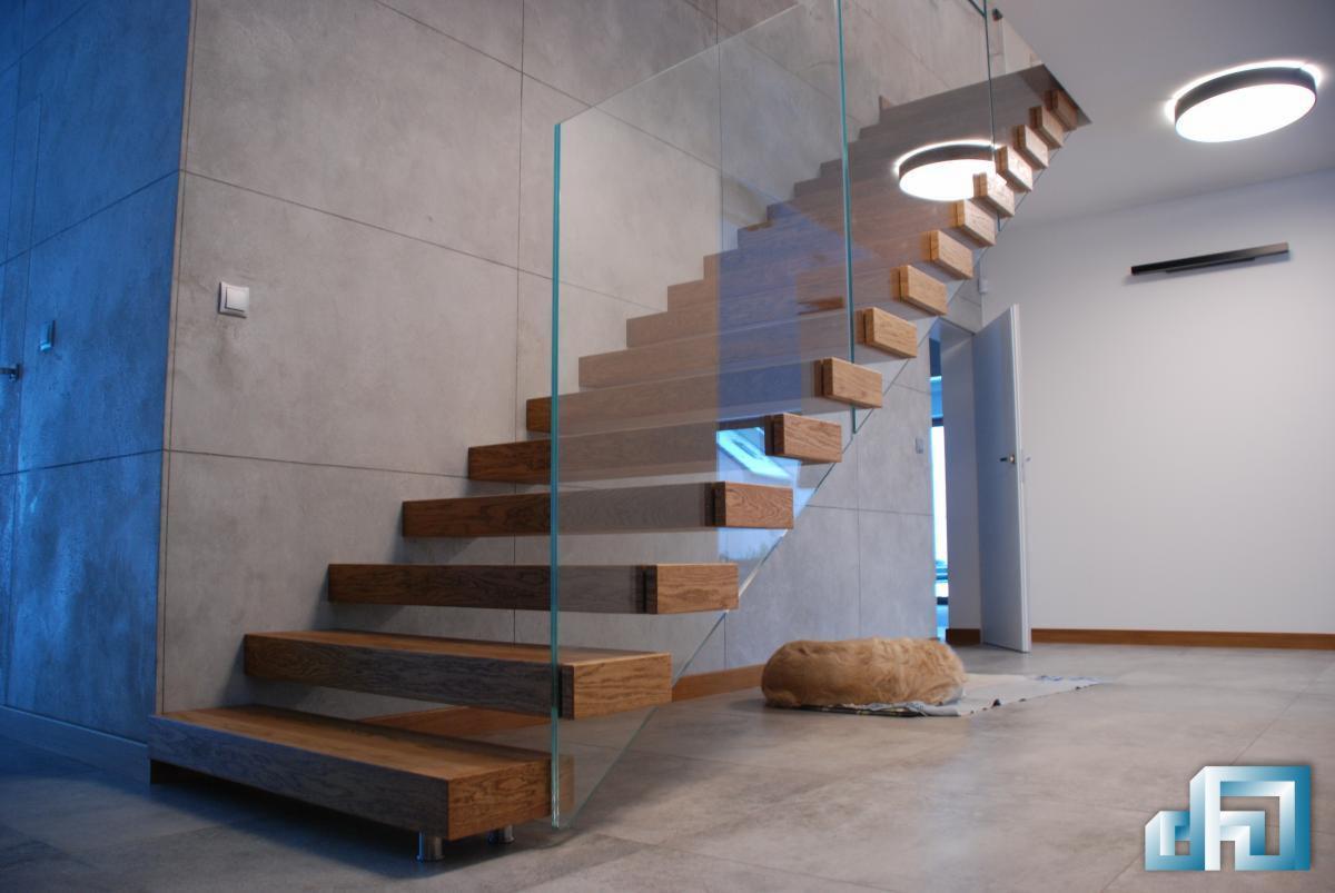 Nowoczesne schody z drewnianymi stopniami i szklaną balustradą, oświetlone okrągłymi lampami sufitowymi, z widocznym fragmentem szarej ściany wykończonej płytkami imitującymi beton i psem leżącym...