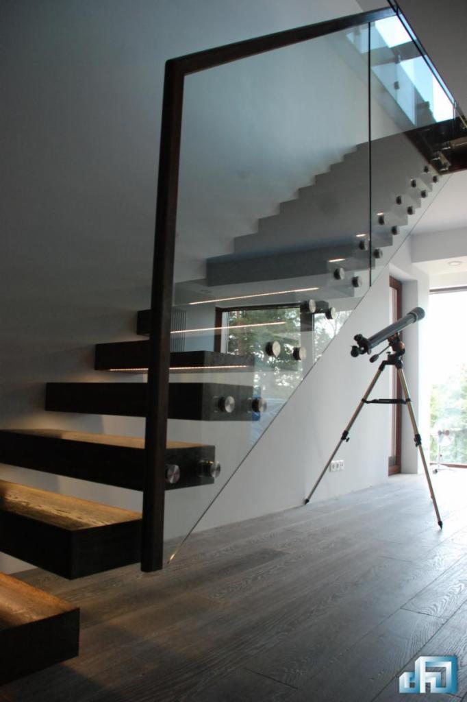 Nowoczesne schody z drewnianymi stopniami i szklaną balustradą, mocowaną za pomocą metalowych uchwytów; na podłodze widoczny teleskop.