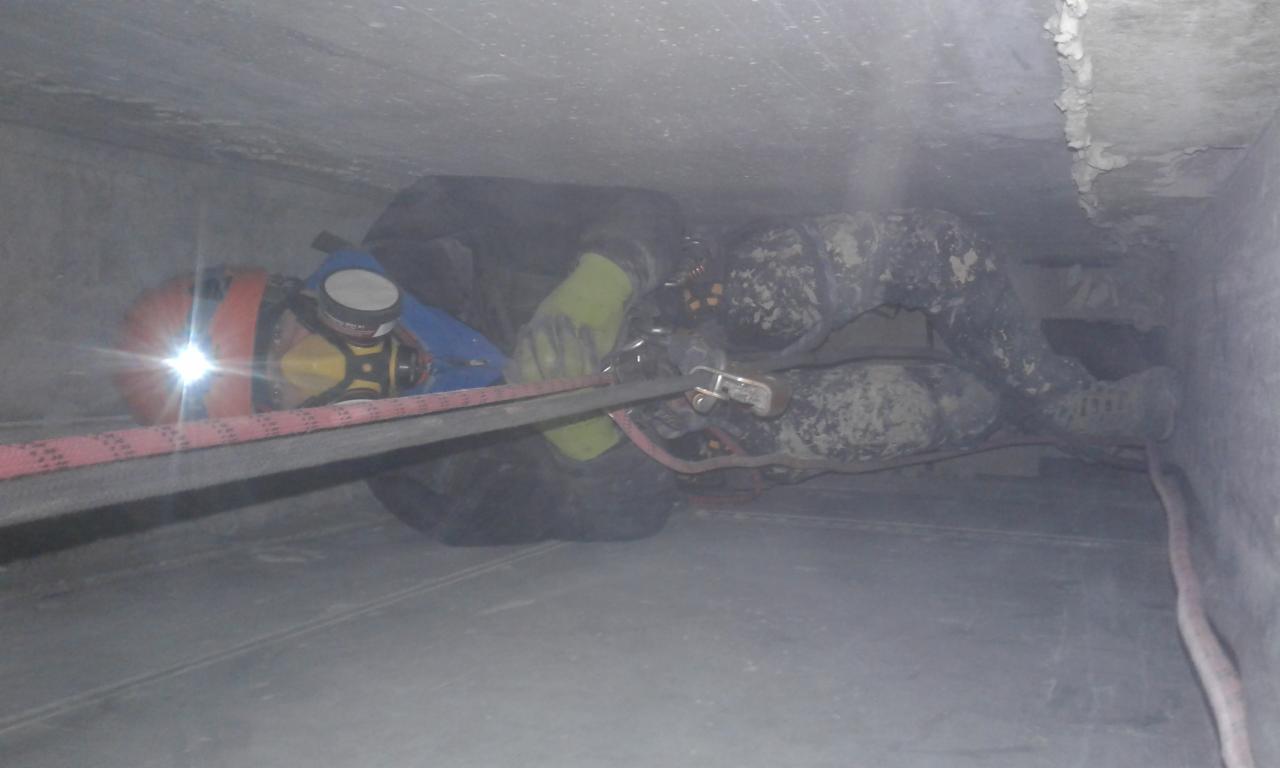 Pracownik w kasku z latarką i masce, zabezpieczony linami, wykonuje prace na wysokości w ciasnej przestrzeni, prawdopodobnie kanał wentylacyjny lub komin