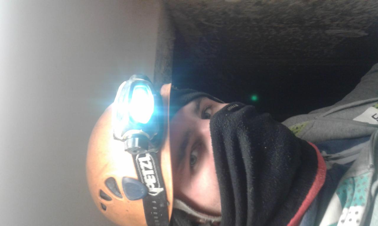 Osoba w kasku Petzl z latarką czołową, częściowo zasłonięta czarną chustą, w ciemnym pomieszczeniu.