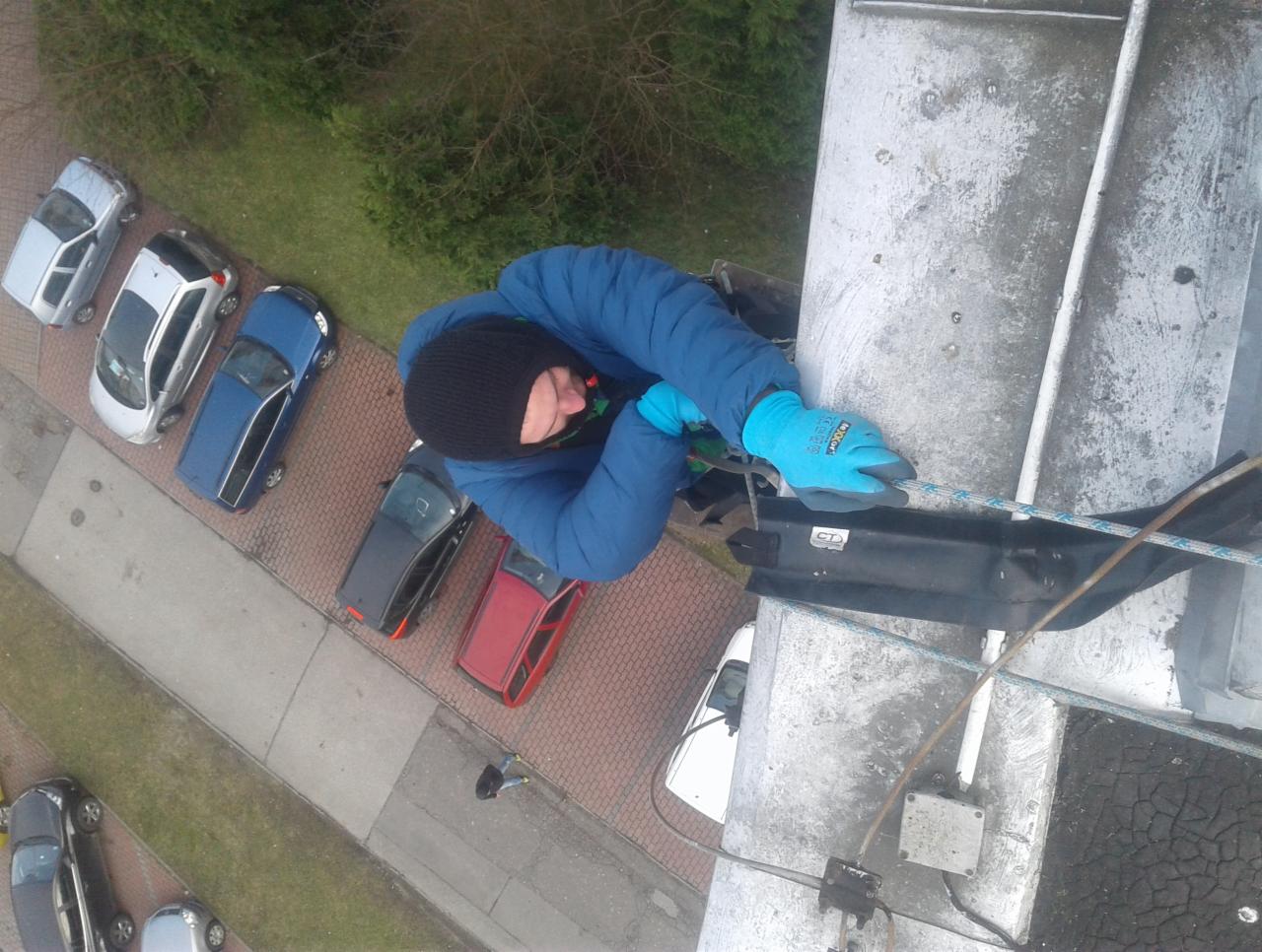 Pracownik w niebieskiej kurtce i rękawicach, z czapką na głowie, zabezpieczony liną, wykonuje prace na wysokości na metalowej konstrukcji, widok z góry na zaparkowane samochody i chodnik z kostki.