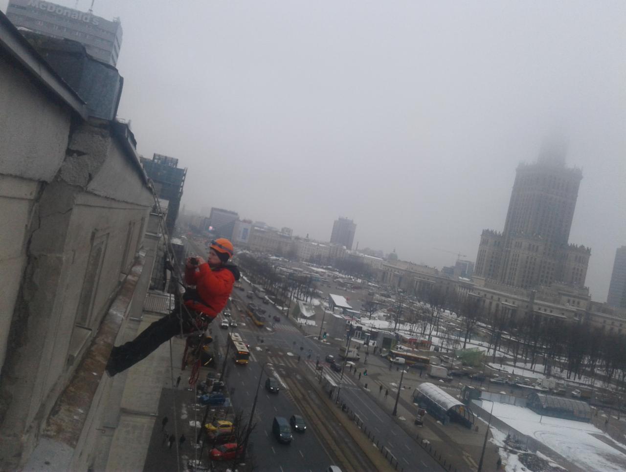 Pracownik na linie zabezpieczającej, w kasku i pomarańczowej kurtce, wykonuje prace na wysokości na elewacji budynku w Warszawie, z widokiem na Pałac Kultury i Nauki w tle, podczas pochmurnego dnia.