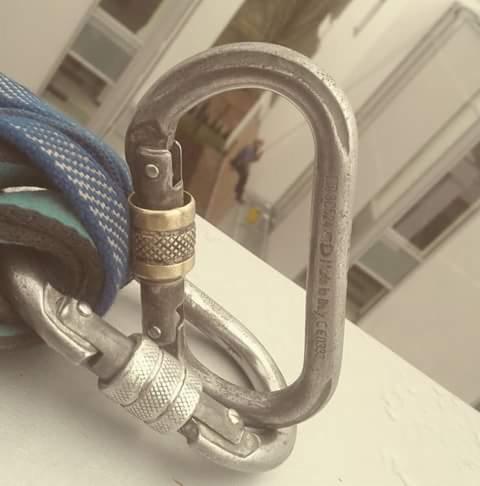 Zapięcie karabińczyka alpinistycznego z liną, w tle pracownik na wysokościach przy elewacji budynku.