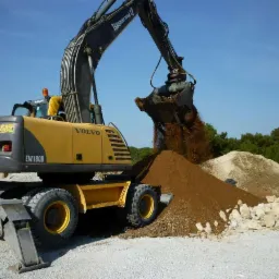 Żółto-szara koparka kołowa Volvo EW180B wysypuje ziemię z łyżki na pryzmę ziemi, w tle widać drzewa i opony.