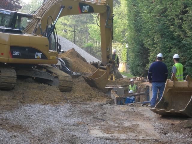 Żółta koparka CAT 320D podczas wykopu ziemi pod fundamenty, widoczni pracownicy w kaskach i kamizelkach odblaskowych, teren budowy z gruzem i ziemią.