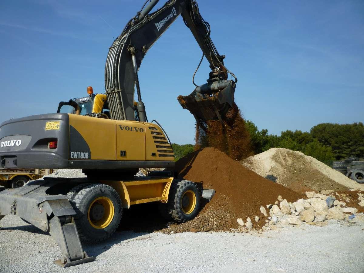 Żółto-szara koparka kołowa Volvo EW180B wysypuje ziemię z łyżki na pryzmę ziemi, w tle widać drzewa i opony.
