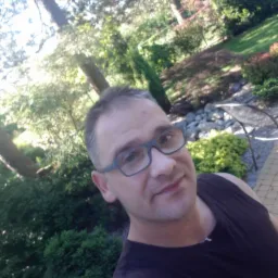 Mężczyzna w okularach na tle ogrodu z drzewami i krzewami. Widoczny fragment ścieżki z kostki brukowej i metalowe krzesło.