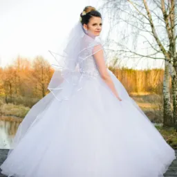 Panna młoda w białej sukni z tiulowym dołem i welonem, spogląda przez ramię na drewnianym pomoście nad wodą, w tle drzewa i zachodzące słońce.