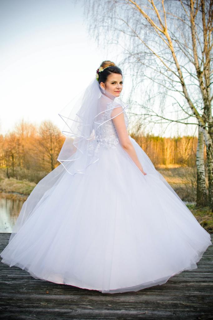 Panna młoda w białej sukni z tiulowym dołem i welonem, spogląda przez ramię na drewnianym pomoście nad wodą, w tle drzewa i zachodzące słońce.