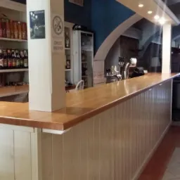 Długi, drewniany blat barowy z białym panelem frontowym w lokalu gastronomicznym, widoczne dystrybutory do piwa, półki z napojami i stoliki w tle.