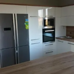 Nowoczesna kuchnia z białymi, lakierowanymi szafkami, drewnianym blatem i szarą lodówką side-by-side, widoczny fragment wyspy kuchennej z blatem drewnopodobnym.