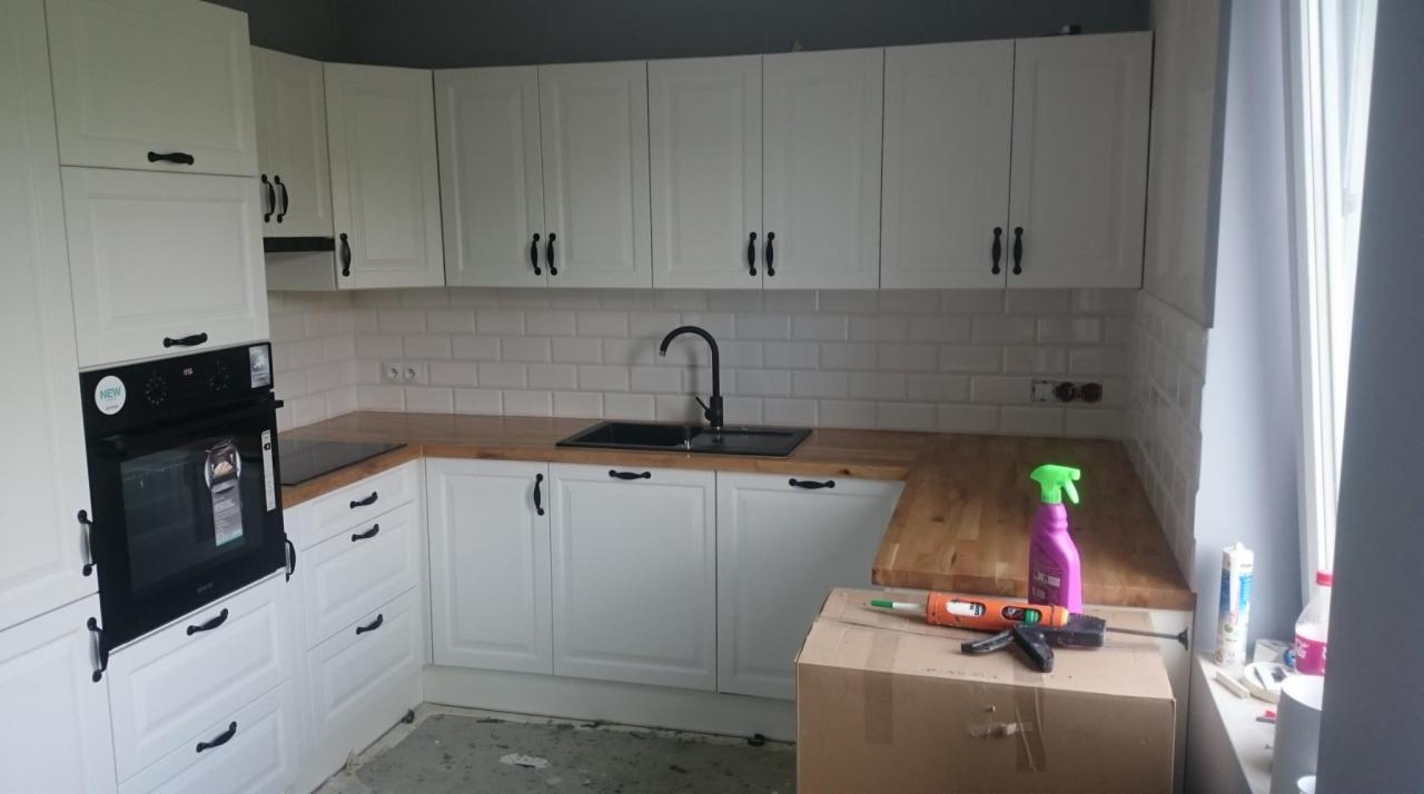 Biała zabudowa kuchenna z drewnianym blatem, czarnymi uchwytami i zlewozmywakiem, widoczna w trakcie montażu, z narzędziami i środkami czyszczącymi na blacie i kartonie.