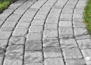 Ścieżka wyłożona szarą kostką brukową ułożoną w łukowaty wzór, widok z góry, obrzeże z trawy po prawej stronie kadru.