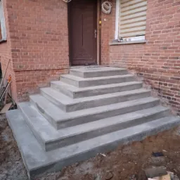 Świeżo wylane betonowe schody prowadzące do drzwi wejściowych budynku z czerwonej cegły, widoczne przewody elektryczne na elewacji.