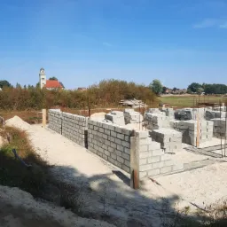 Wznoszenie fundamentów budynku z bloczków betonowych na piaszczystym terenie, widoczna konstrukcja zbrojeniowa, w tle kościół z wieżą i błękitne niebo.
