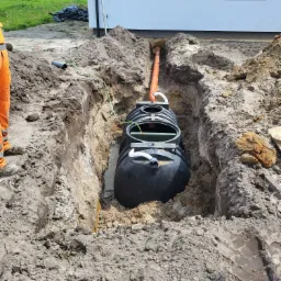 Czarny zbiornik bezodpływowy w wykopie przy budynku, widoczne rury kanalizacyjne i kabel elektryczny, częściowo zasypany ziemią.