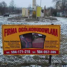 Żółty baner reklamowy firmy ogólnobudowlanej z numerami telefonów na tle pozostałości budynku po rozbiórce, widoczny komin i śnieg.