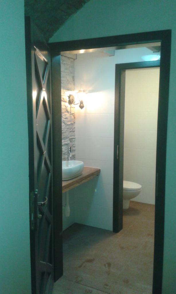 Otwarta, ciemna, metalowa rama drzwi prowadzi do wyremontowanej łazienki z białą umywalką nablatową na drewnianym blacie, kamienną ścianą i toaletą w tle.