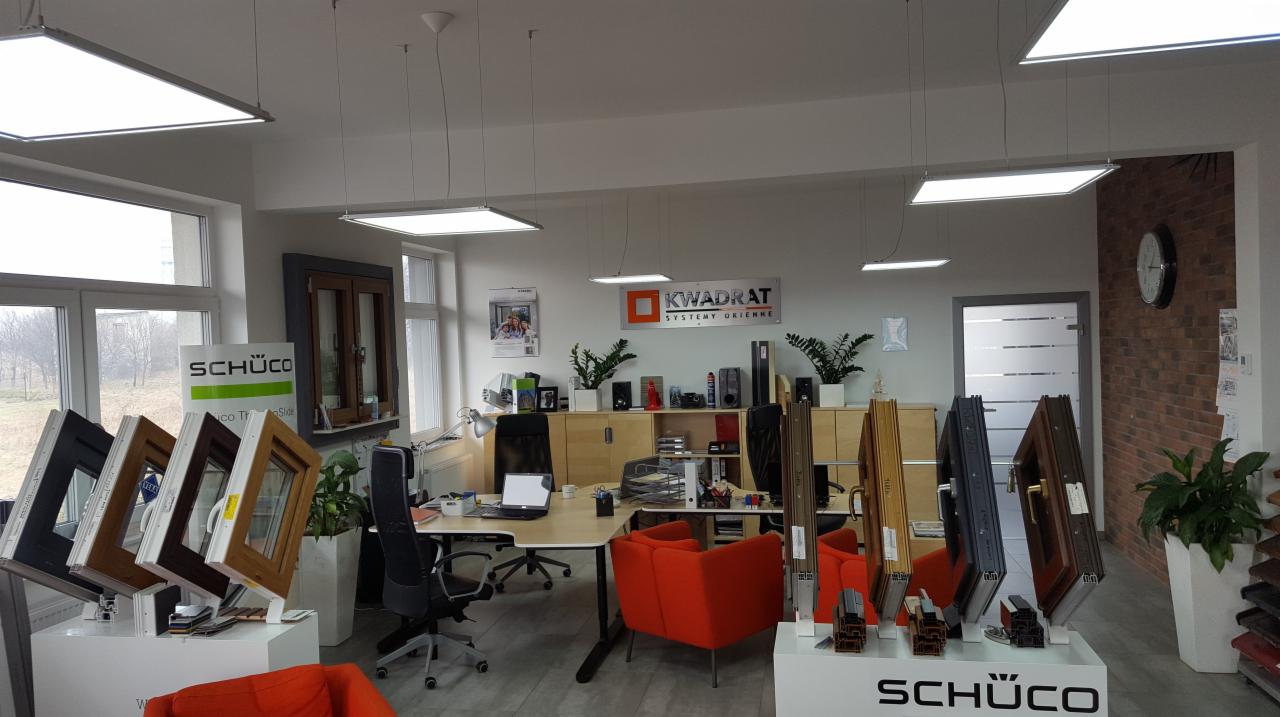 Wnętrze biura firmy oferującej okna i drzwi, ekspozycja profili okiennych i drzwiowych marki Schüco, widoczne biurka, krzesła, rośliny doniczkowe oraz logo firmy Kvadrat Systemy Okienne.