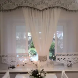 Eleganckie, białe, transparentne firany z dekoracyjnym, wzorzystym lambrekinem i ozdobnym wykończeniem, widok na okno z dekoracjami świątecznymi na parapecie i ogrodem w tle.