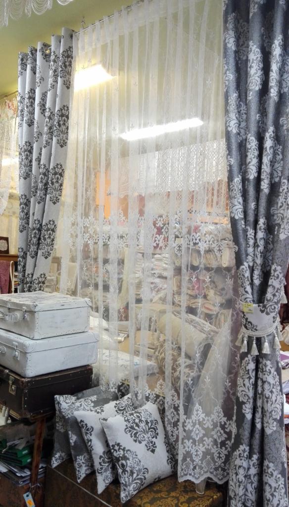 Aranżacja okienna z wykorzystaniem białych, koronkowych firan i zasłon w odcieniach szarości z motywem roślinnym, uzupełniona dekoracyjnymi poduszkami i walizkami.