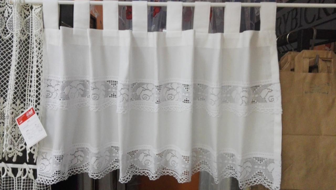 Krótka, biała zasłonka z trzema rzędami koronkowych wstawek, zawieszona na karniszu za pomocą białych pasków, obok widoczna koronkowa serwetka z czerwoną metką.