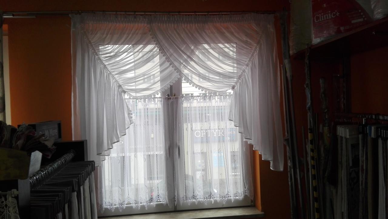 Białe, gotowe firany z woalu i gipiury zdobią okno w Zambrzyńcu, widoczne zza nich szyld optyka.