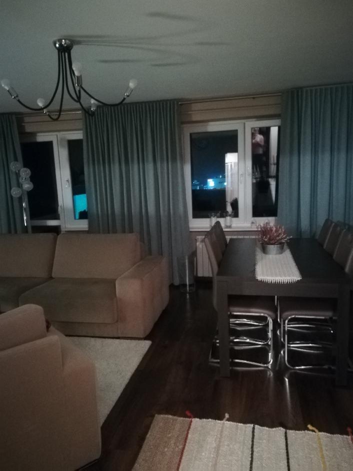 Salon z beżową sofą i ciemnym stołem jadalnym, okna zasłonięte turkusowymi, długimi zasłonami, widok na nocne miasto.