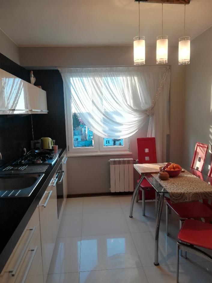 Jasna kuchnia z białymi meblami i oknem przesłoniętym białą, zwiewną zasłoną z dekoracyjnymi koralikami, wisząca lampa z trzema szklanymi kloszami nad stołem z owocami.