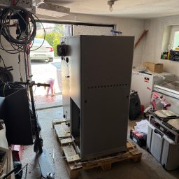 Pro-Weld Sp. z o.o. - Duża, szara centrala wentylacyjna na palecie w garażu, widoczne rury wentylacyjne i sprzęt. Dziecko bawi się na zewnątrz, widoczne przez otwarte drzwi garażowe.