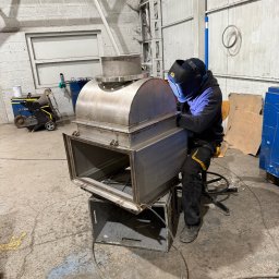 Pro-Weld Sp. z o.o. - Spawacz w masce spawalniczej łączy metalowe elementy konstrukcji wentylacyjnej w hali, używając spawarki. Widoczne iskry i elementy konstrukcyjne.