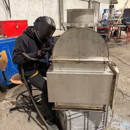 Pro-Weld Sp. z o.o. - Spawacz w czarnym kombinezonie i masce spawalniczej łączy metalowe elementy konstrukcji w hali przemysłowej. Widoczne iskry i spawarka Fronius.