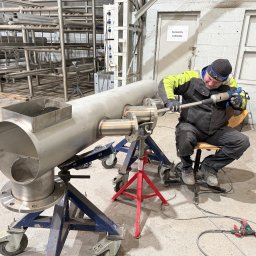 Pro-Weld Sp. z o.o. - Spawanie rury ze stali nierdzewnej przez montera w warsztacie. Rura na wózku warsztatowym. Widoczne narzędzia i wyposażenie. Magazyn Carbone w tle.