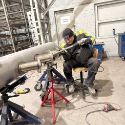 Pro-Weld Sp. z o.o. - Spawacz w Szczytnie łączy rury ze stali nierdzewnej w hali produkcyjnej, używając profesjonalnego sprzętu spawalniczego. Widoczne zabezpieczenia i narzędzia.