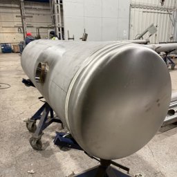 Pro-Weld Sp. z o.o. - Metalowy, cylindryczny zbiornik na niebieskim wózku, z widocznym spawem i otworem. W tle hala produkcyjna z pracownikami i konstrukcjami stalowymi.