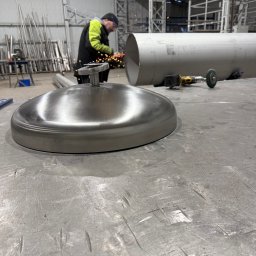 Pro-Weld Sp. z o.o. - Spawacz w hali obrabia metalową rurę szlifierką kątową. Na pierwszym planie polerowana metalowa pokrywa. W tle rury i konstrukcja hali.