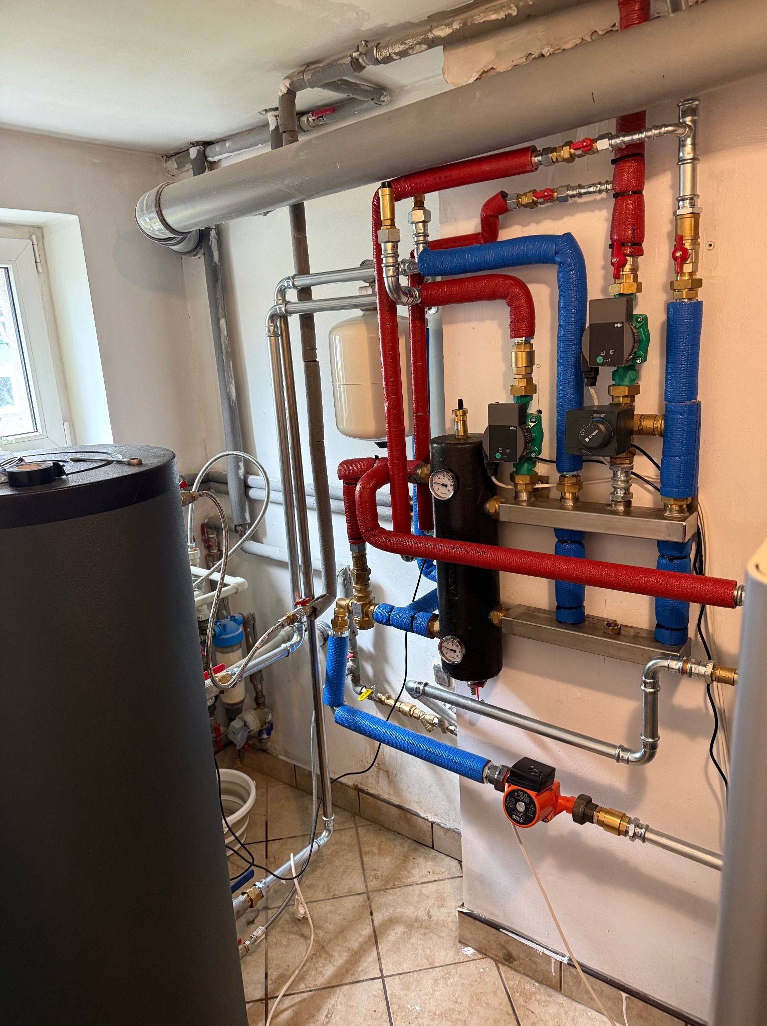 Złożona instalacja grzewcza z rurami w izolacji (czerwonej i niebieskiej), zbiornikiem i manometrami. Widoczne liczne zawory i pompy obiegowe. Ujęcie w kotłowni.