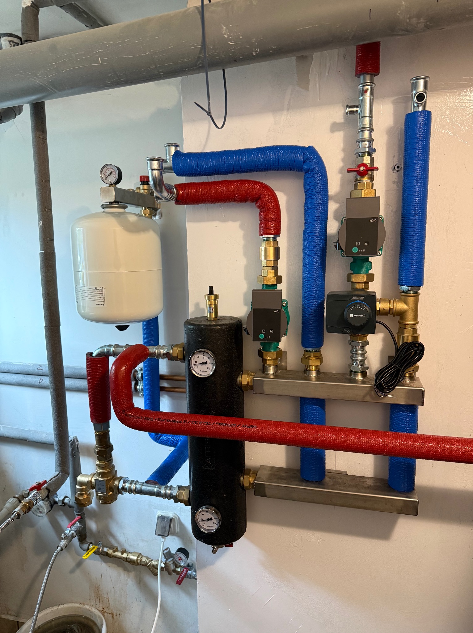 Instalacja CO z zbiornikiem wyrównawczym, rurami w izolacji czerwonej i niebieskiej, manometrami i zaworami, zamontowana na ścianie w kotłowni.
