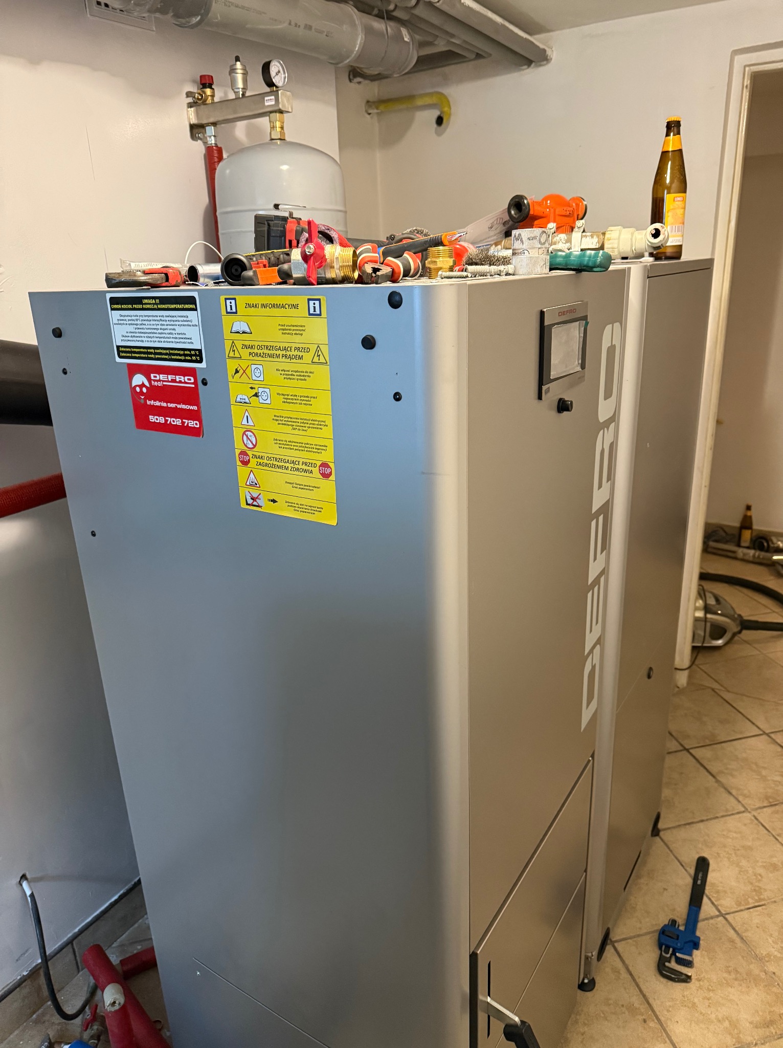 Szary piec Defro z narzędziami i akcesoriami na górze w pomieszczeniu technicznym. Widoczny manometr, rury i ostrzeżenia. Na podłodze płytki.