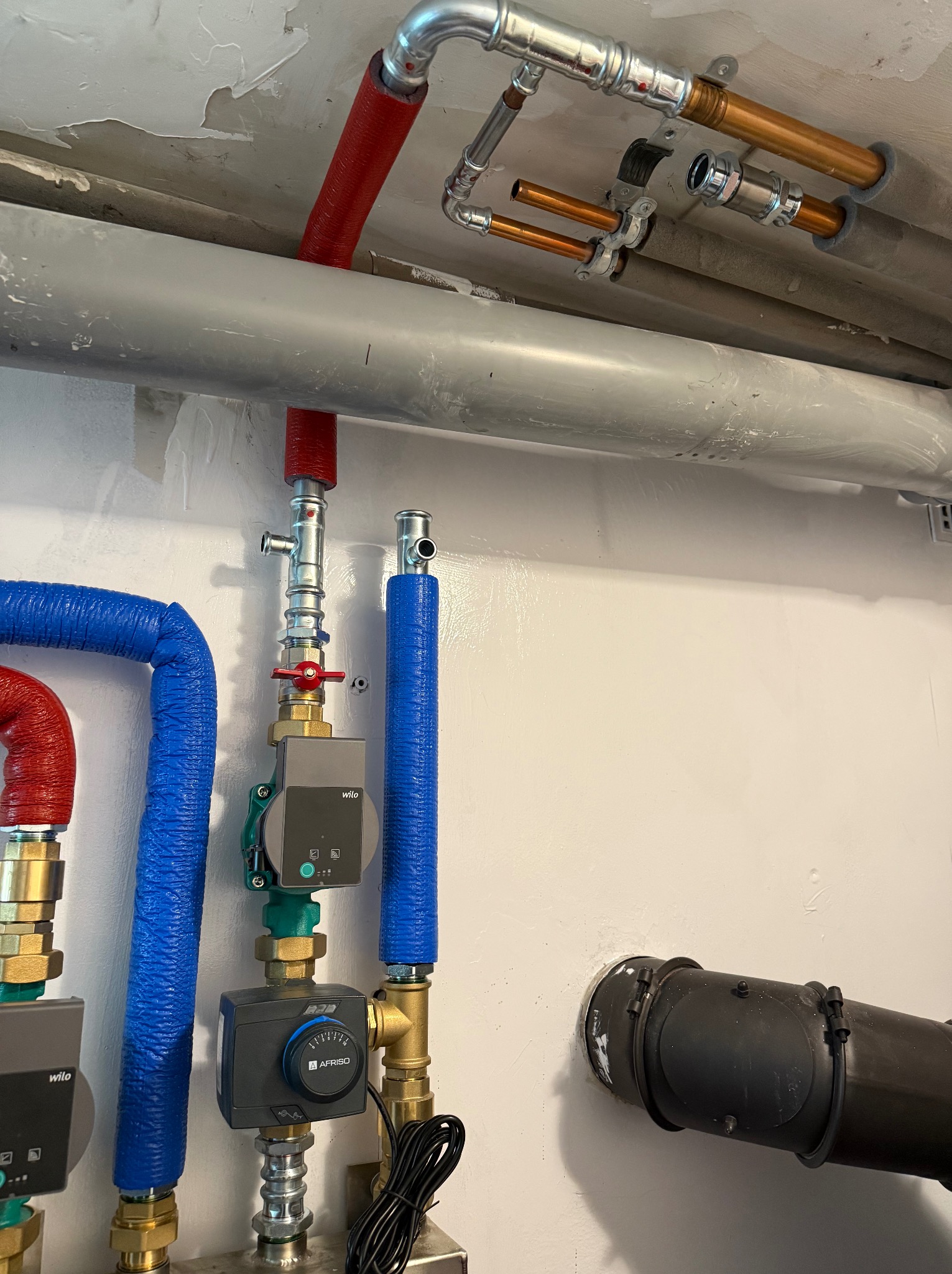 Instalacja hydrauliczna z rurami miedzianymi i stalowymi, izolowanymi na czerwono i niebiesko, z zaworami i pompami obiegowymi Wilo, zamontowana przy suficie.