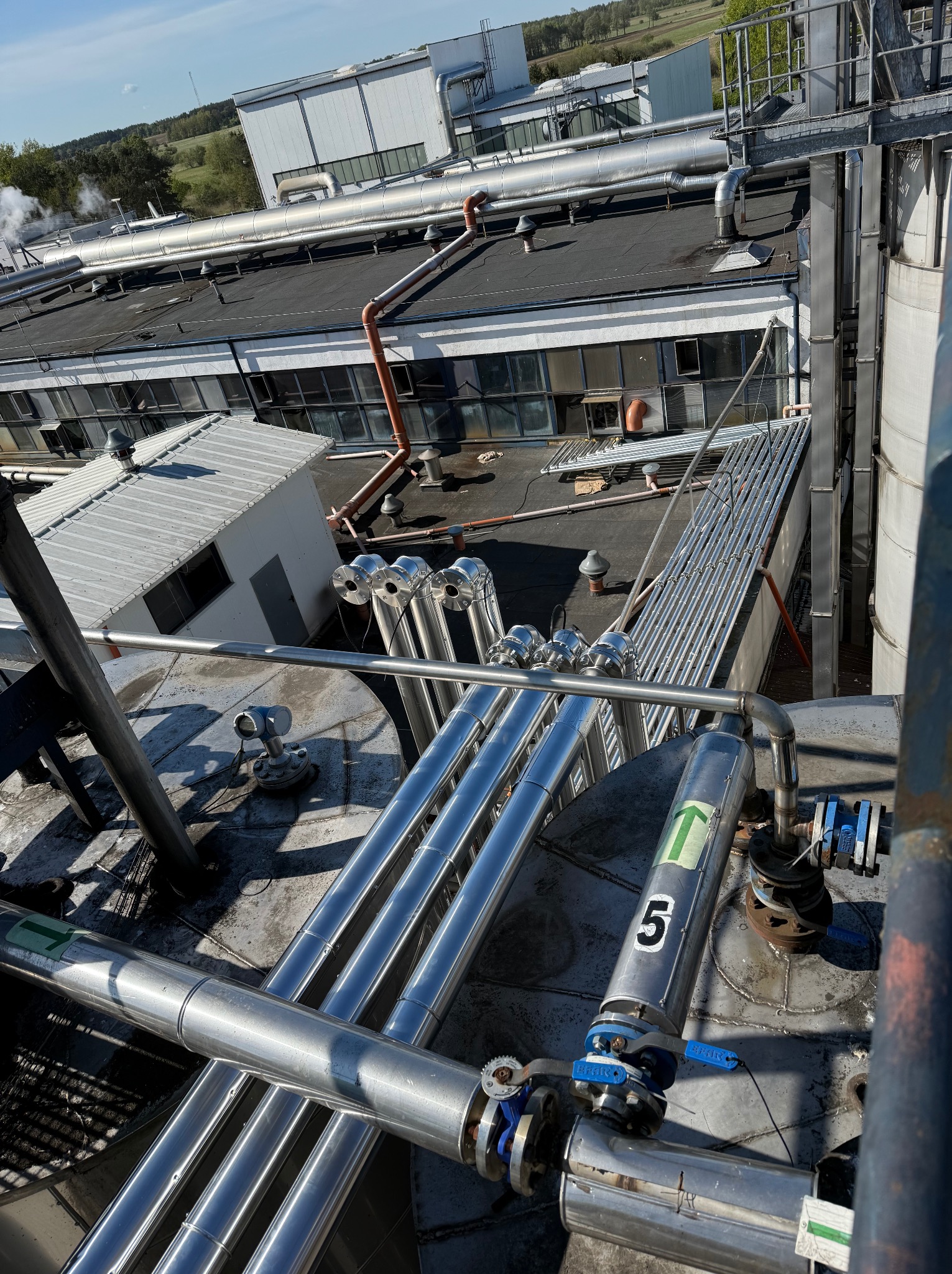 Widok z góry na przemysłowe instalacje rurociągowe na dachu budynku fabrycznego; rury ze stali nierdzewnej z zaworami i oznaczeniami, tło stanowią inne budynki i zieleń.