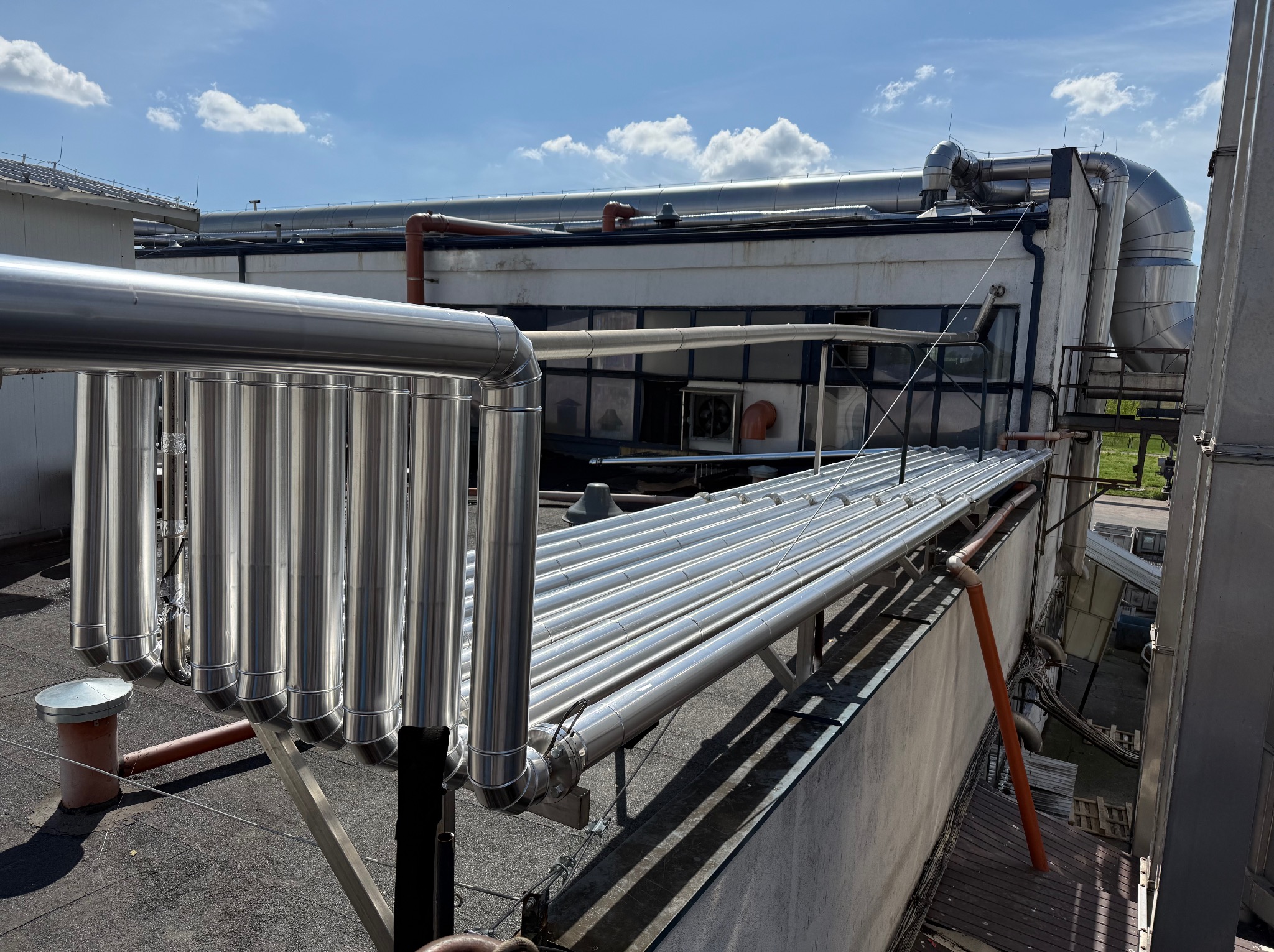 Instalacja przemysłowa na dachu budynku z licznymi rurami ze stali nierdzewnej, widoczne elementy wentylacji i orurowania, jasne niebo z chmurami w tle.