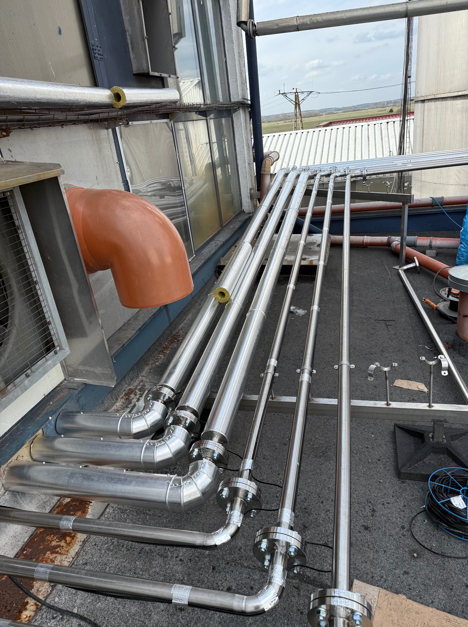Instalacja przemysłowa: chromowane rury z kołnierzami, pomarańczowy element wentylacyjny, widok na zewnątrz budynku, instalacje na dachu.