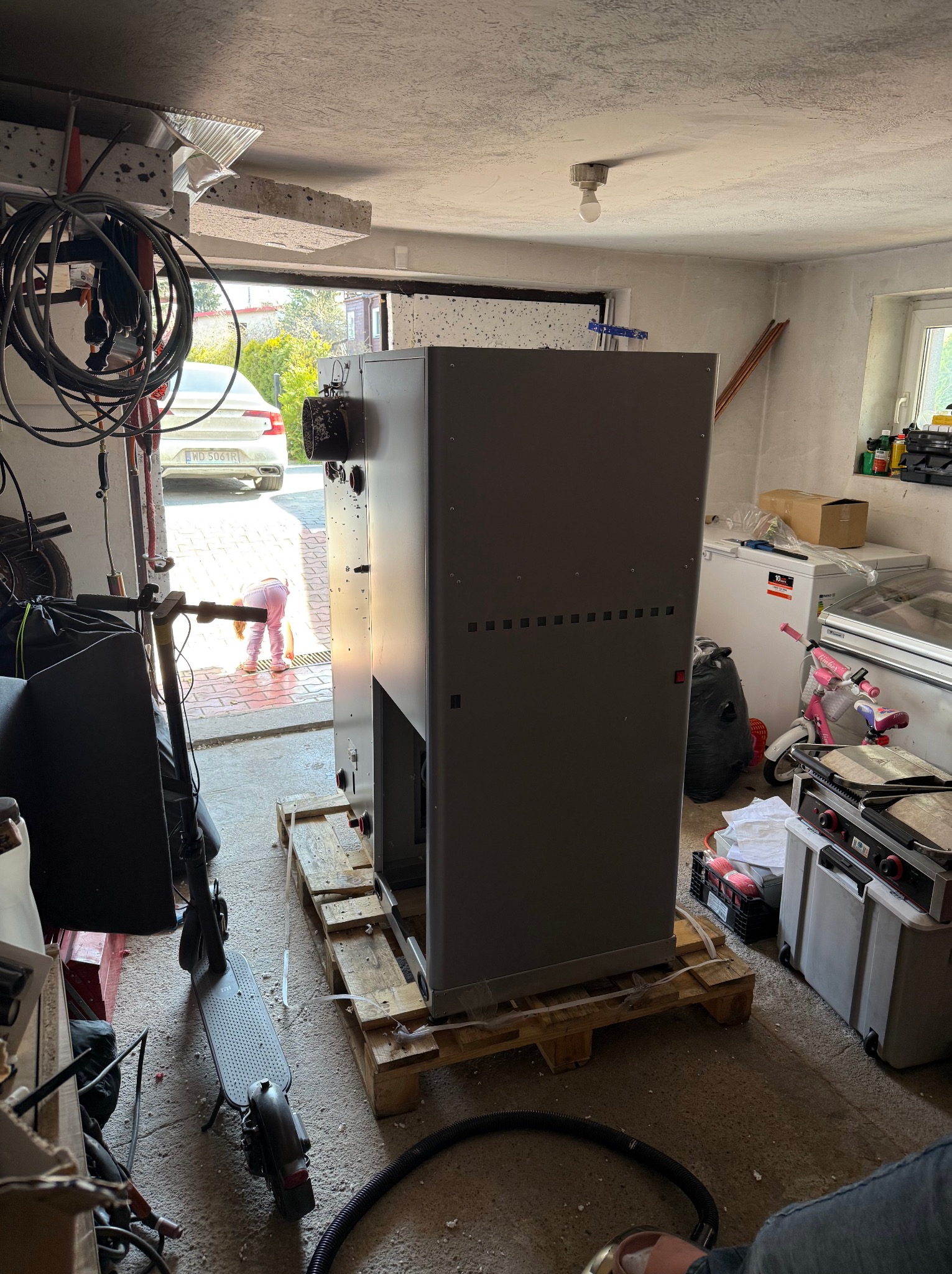 Duża, szara centrala wentylacyjna na palecie w garażu, widoczne rury wentylacyjne i sprzęt. Dziecko bawi się na zewnątrz, widoczne przez otwarte drzwi garażowe.