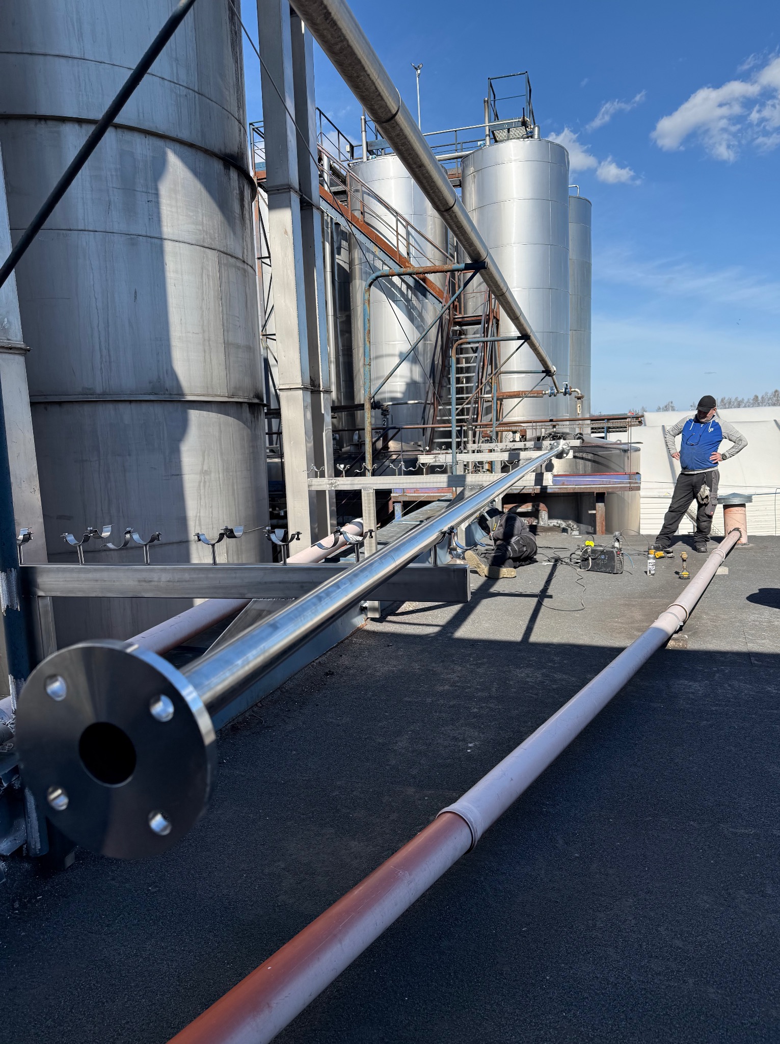 Montaż rurociągu przemysłowego na dachu, spawanie rur, widoczne zbiorniki i drabiny. Dwóch monterów przy pracy, narzędzia na dachu. Instalacje przemysłowe w słoneczny dzień.