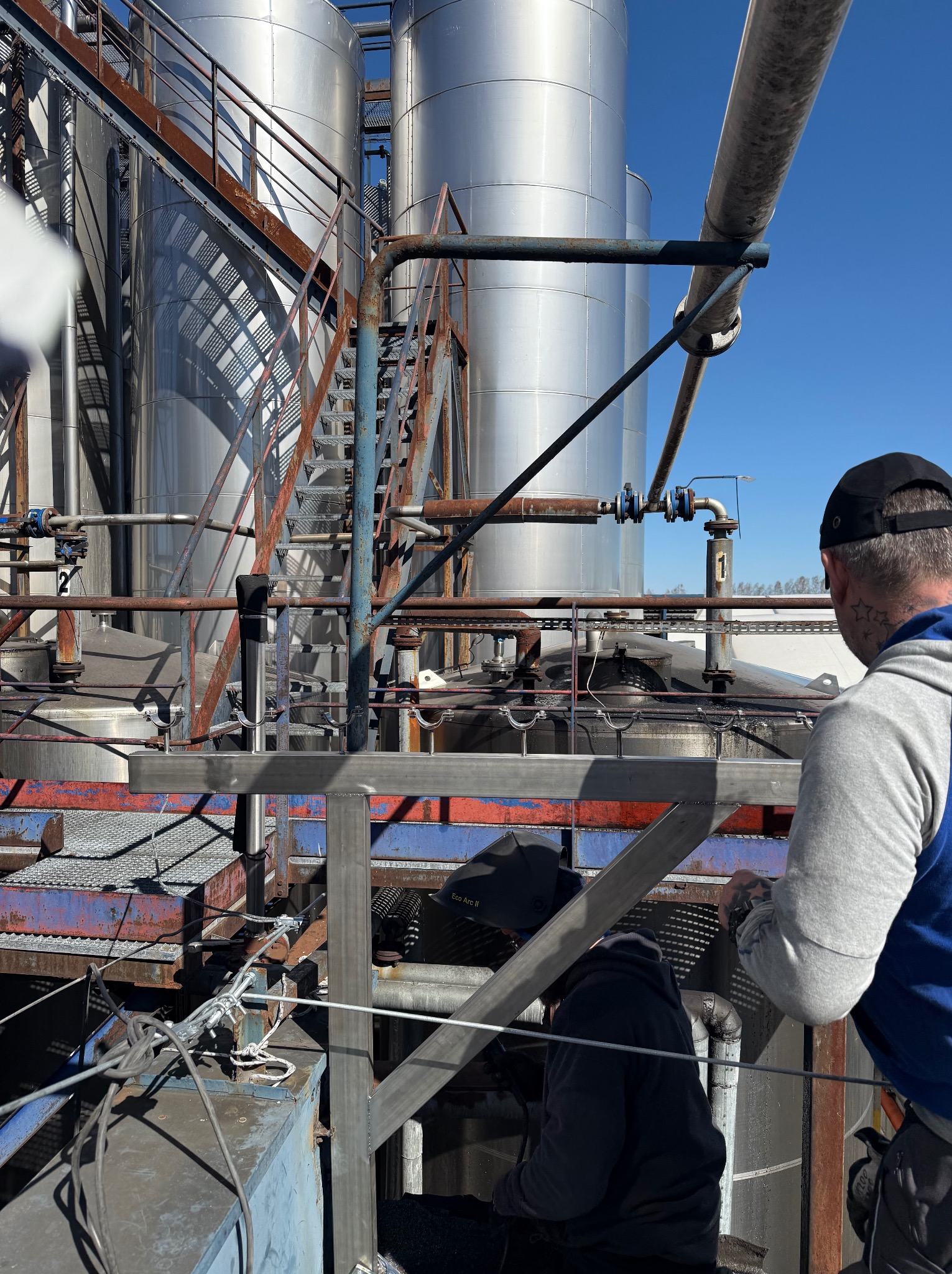 Prace montażowe na instalacji przemysłowej: spawanie rurociągów przy zbiornikach, widoczni dwaj monterzy w trakcie pracy, niebieskie niebo w tle.