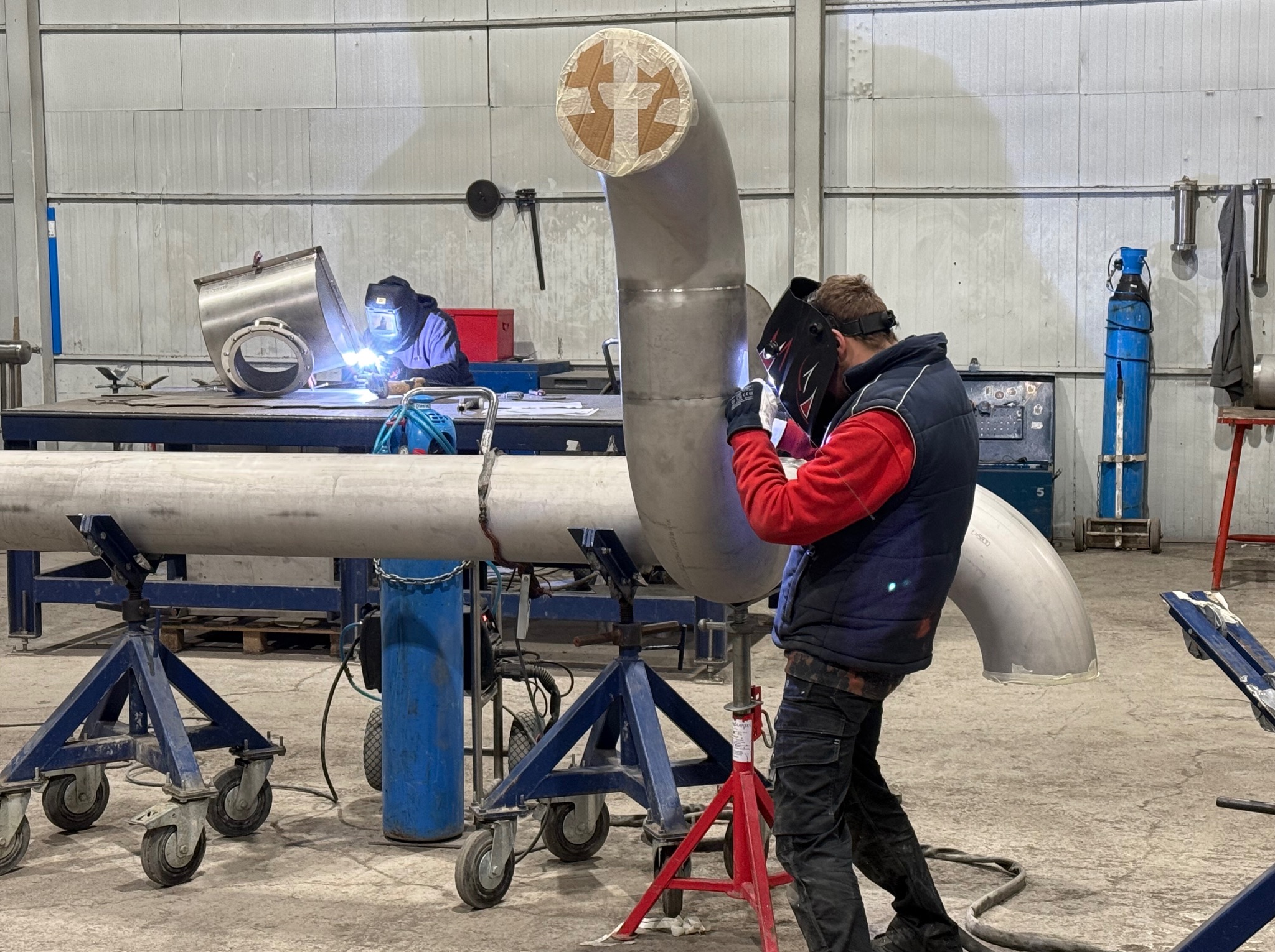 Spawanie rur ze stali nierdzewnej w hali produkcyjnej. Spawacz w masce spawalniczej łączy elementy rurociągu. W tle drugi spawacz przy pracy.