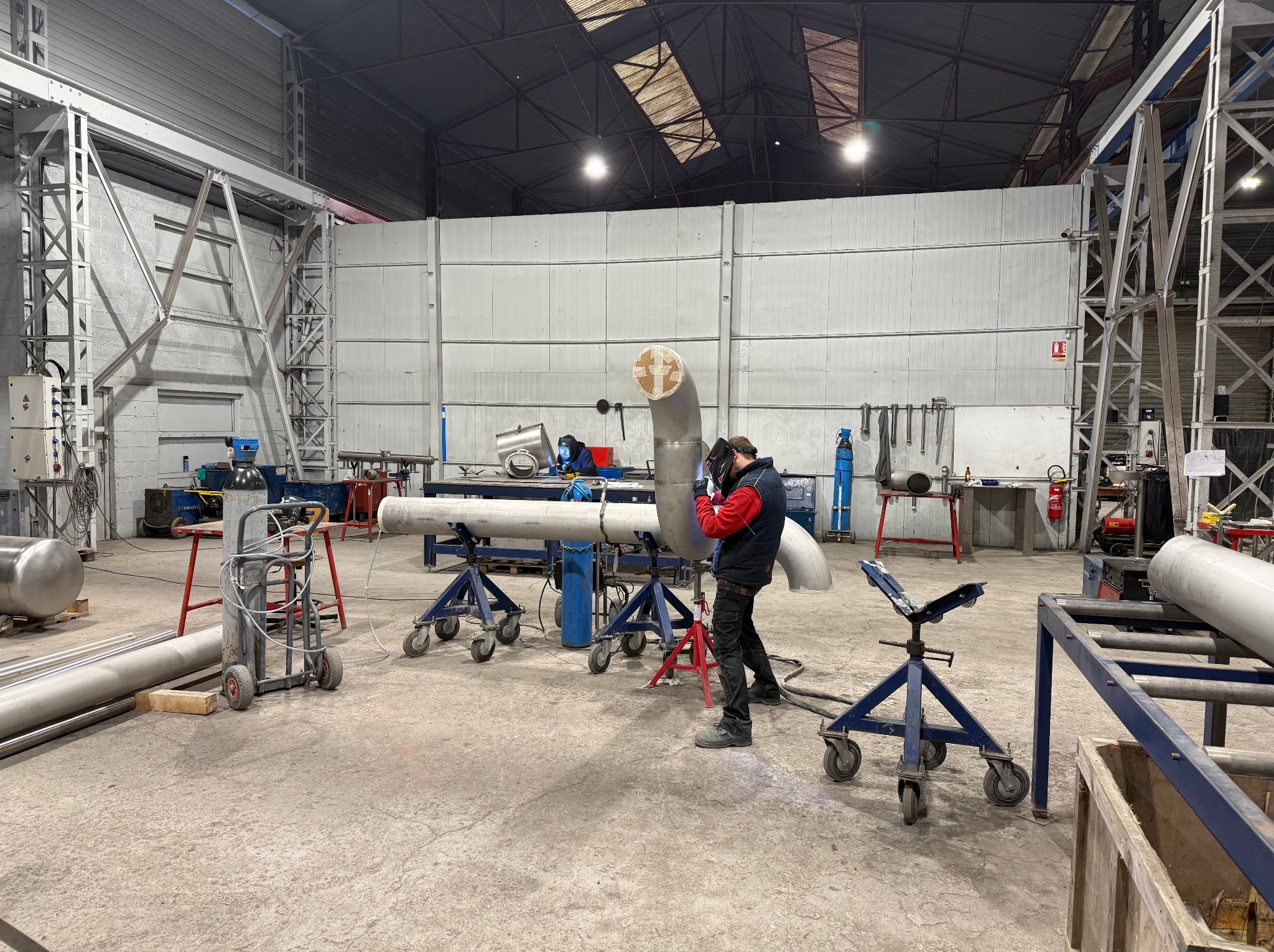 Spawanie rur w hali produkcyjnej. Pracownik w kombinezonie spawalniczym łączy elementy rurociągu. Widoczne narzędzia, butle z gazem i konstrukcja hali.