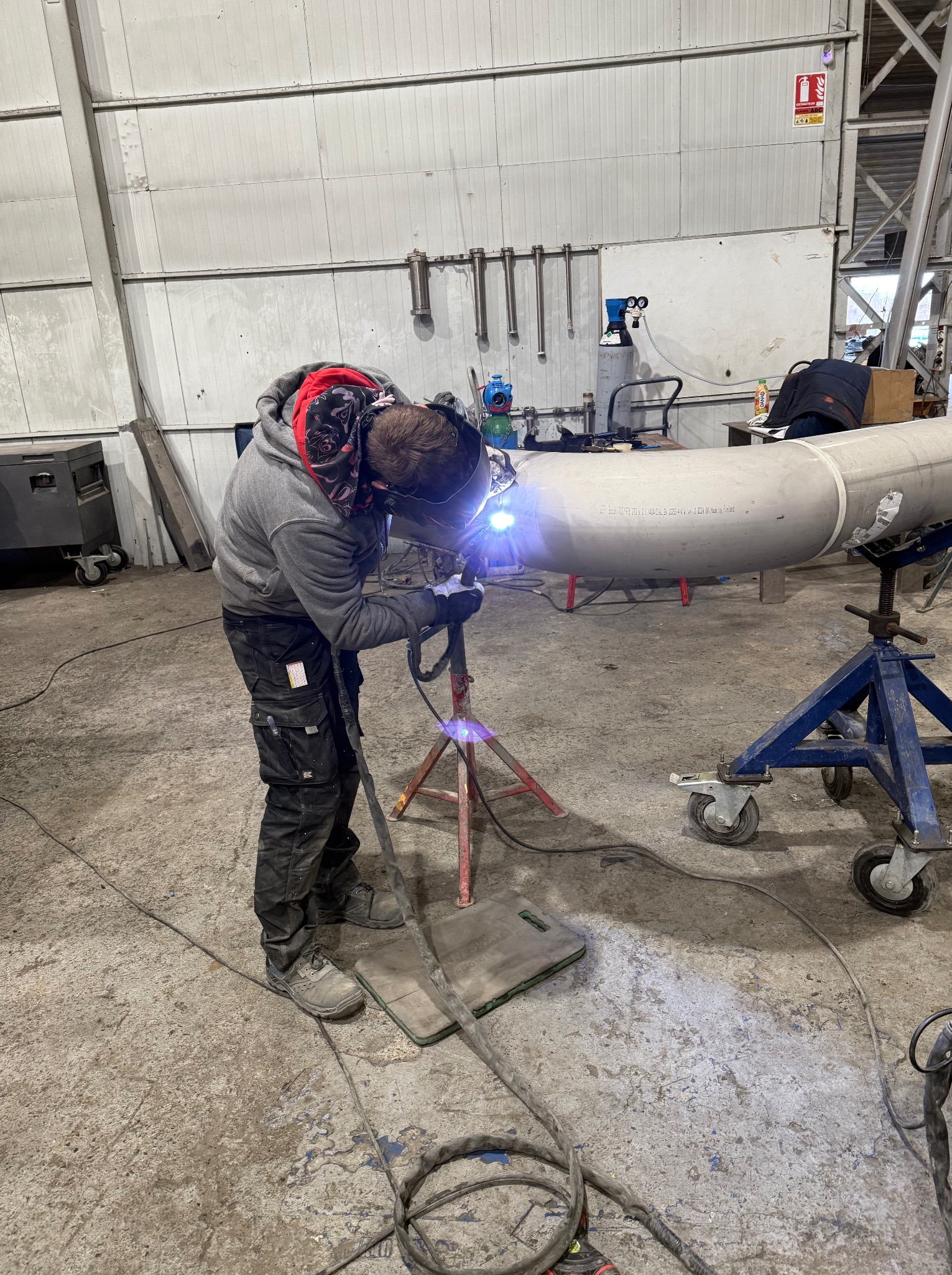 Spawacz w masce spawalniczej łączy rurę na stojaku w hali. Widoczne narzędzia i iskry. Rura z napisem: Made in Finland.